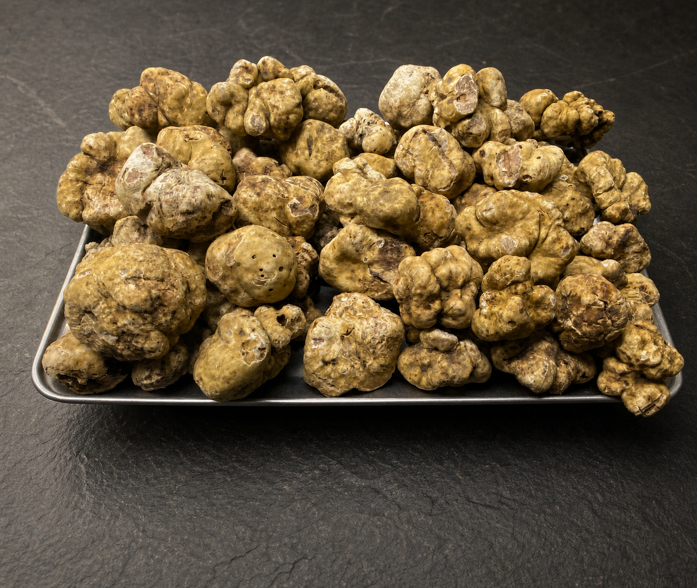 A large tray of irregular fresh white truffles on a dark slate surface