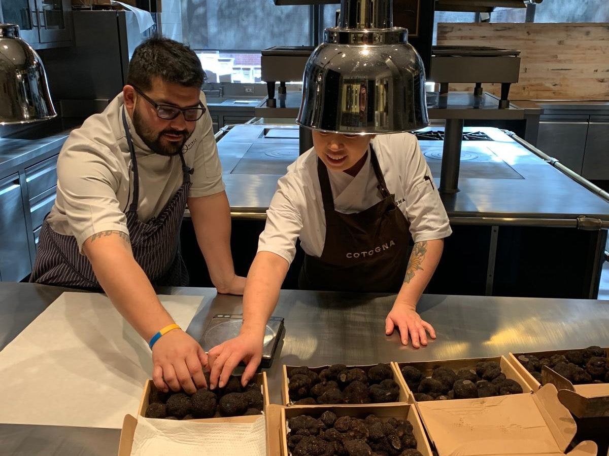 Two chefs inspect and weigh fresh black truffles beside open truffle boxes in a professional kitchen.